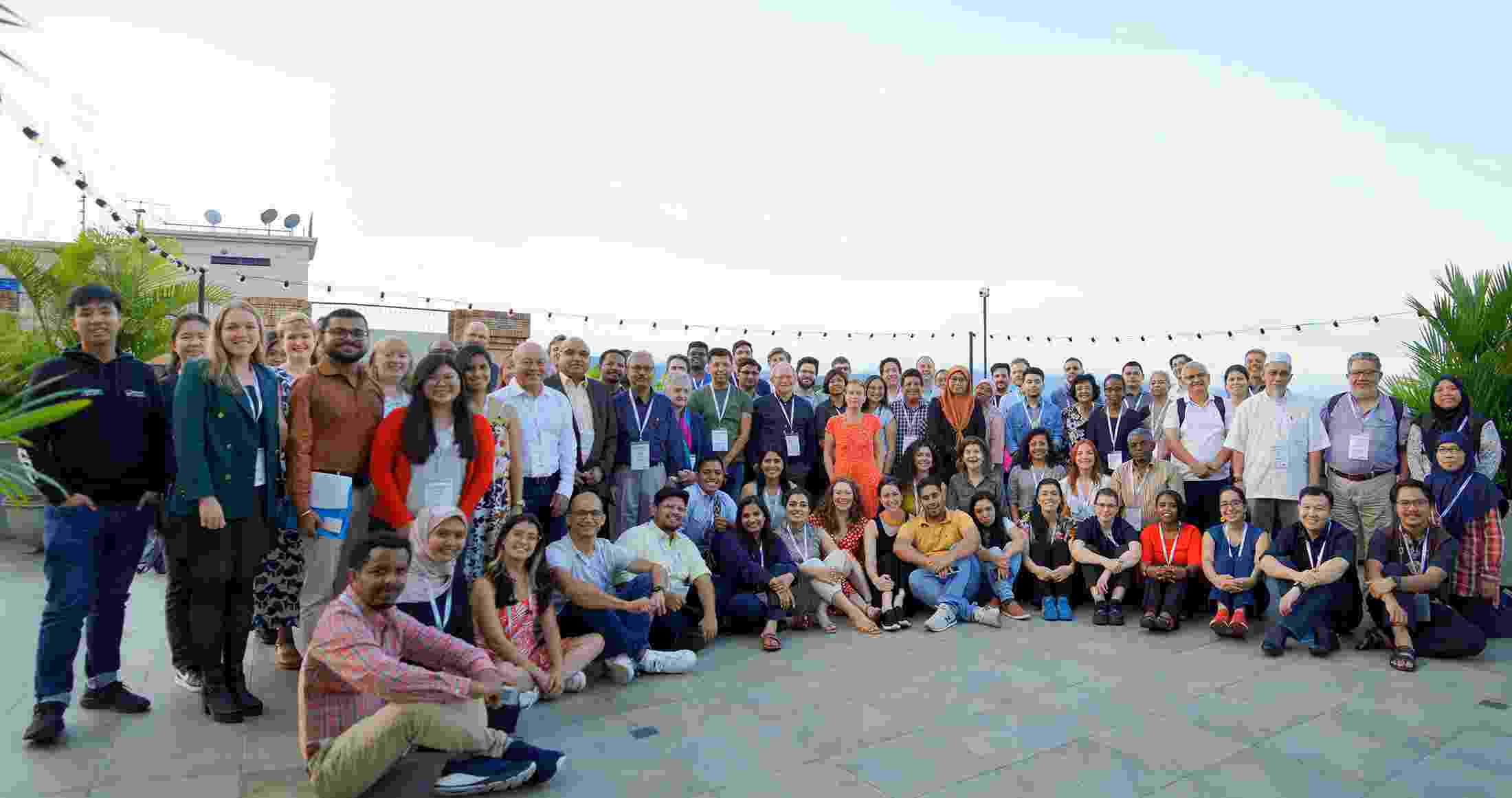 A large mixed group of Hub members sit and stand together in several rows, smiling at the camera, with rooftops and blue sky visible behind them